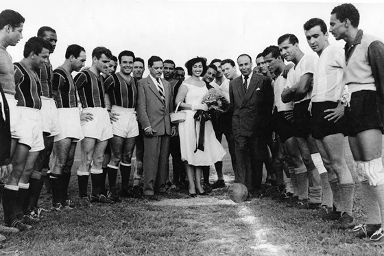 Saque de honor del gobernador de Norte de Santander Gonzalo Rivera Laguado en un partido de fútbol del Cúcuta Deportivo en 1955. A la derecha la madrina del equipo y el Doctor Luís Figueredo Corrales, rodeados por los jugadores locales y visitantes.